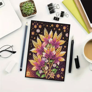 Diamond painting notesbog med blomster i lilla og gyldne toner.